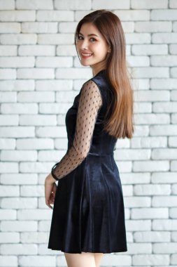 Zarif siyah elbise giyen, beyaz tuğla duvarın arka planında kendine güvenen ve kameraya bakan Asyalı zevkli bir kadın.