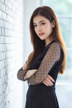 Hoş bir Asyalı kadın portresi, Lao etnik kökenli, şık siyah elbiseli, kollarını kavuşturmuş odada duruyor ve kameraya özgüvenle bakıyor..