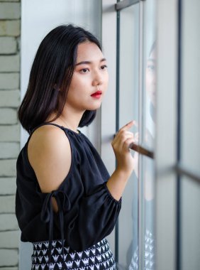 Yakışıklı Asyalı kadın yarı vücut portresi, Lao etnik kökenli, şık siyah elbise içinde, pencerenin yanında duran sevimli ve sakin duruş ve diğer tarafa bakıyor.