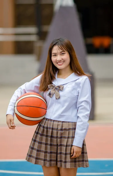 大学キャンパスのスポーツグラウンドでバスケットボールと制服姿の正アジア女性学生とカメラを見る — ストック写真