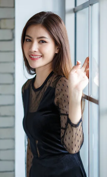 Side view portrait of charming Asian female, Lao ethnicity, in elegant black dress standing with cute and calm pose near window and looking at camera