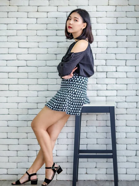 魅力的なアジアの女性でスタイリッシュなスカートとブラウス座っています上のスツール上の白いレンガの壁の背景にスタジオ — ストック写真