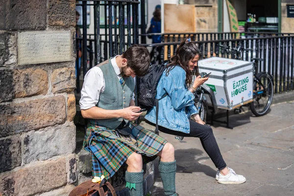 Edinburgh, Skottland, 18 augusti 2019. En ung man klädd i nationella skotska kläder och en kilt använder en smartphone sitter på en trottoarkant — Stockfoto