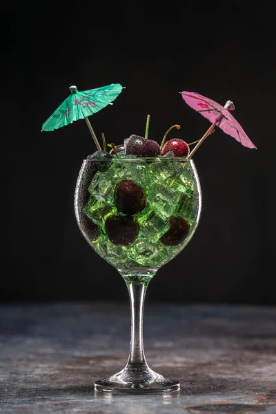 Зеленый коктейль со льдом и вишней в стакане, украшенном зонтиками — стоковое фото