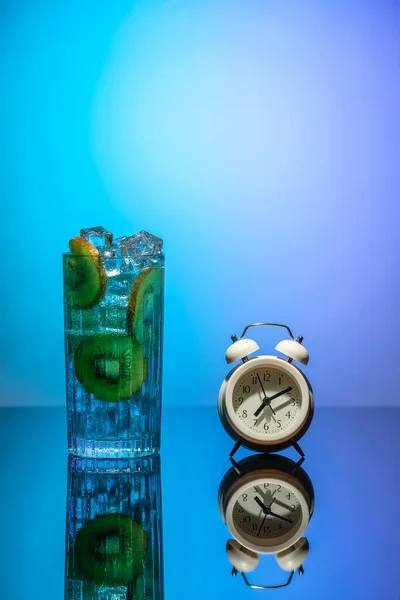 A glass of water with ice and slices of kiwi standing on a mirror table next to the alarm clock. Body detoxification concept