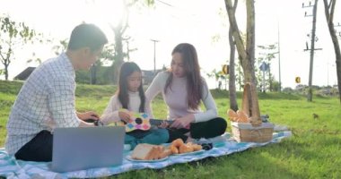 Genç aile parkta oturmuş piknik yapıyor, kızı da küçük gitarı tutuyor.