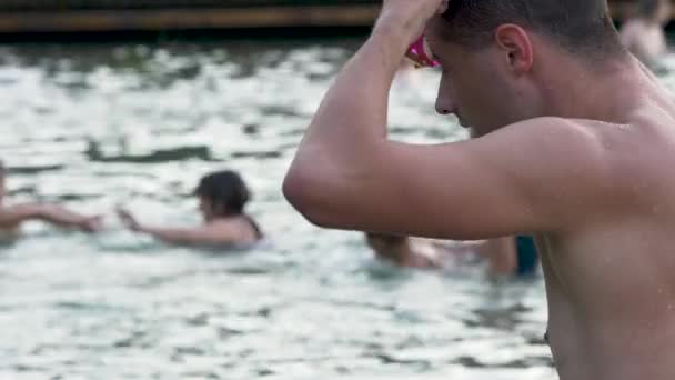 El joven pone anteojos de nadador antes de entrenar. Atleta se prepara para entrenar en el lago. Primer plano de nadador cansado. La gente disfruta del agua tibia en el fondo. — Vídeo de stock