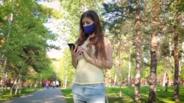 Elinde akıllı telefon olan maskeli bir kız şehir parkında dururken sosyal ağların sayfalarını karıştırıyor..