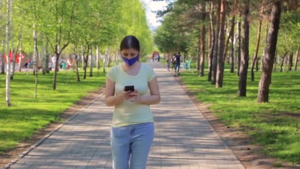 Seorang Wanita Muda Bertopeng Biru Dan Smartphone Tangannya Berjalan Taman — Stok Video