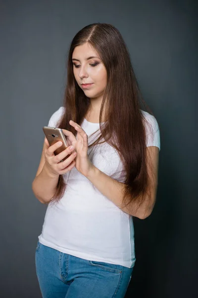 白いTシャツを着た若い美しい女性とスマートフォンを手にした青いジーンズ 灰色の背景のスタジオ写真 — ストック写真