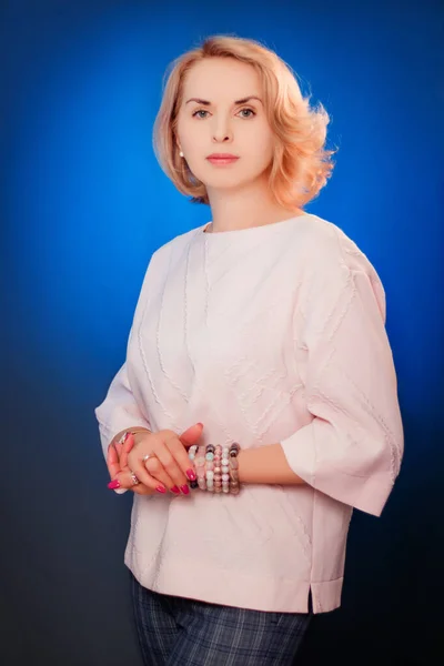 Portrait of an aged woman posing with hands clasped together, standing on blue background.