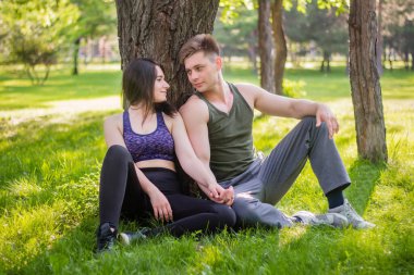 Atletik adam ve kız çimenlerde oturup ağaca yaslanıyorlar..