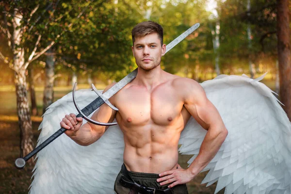 A young angel, a bodybuilder in plate armor on his legs with wings behind his back, holds in his hands a two-handed sword.