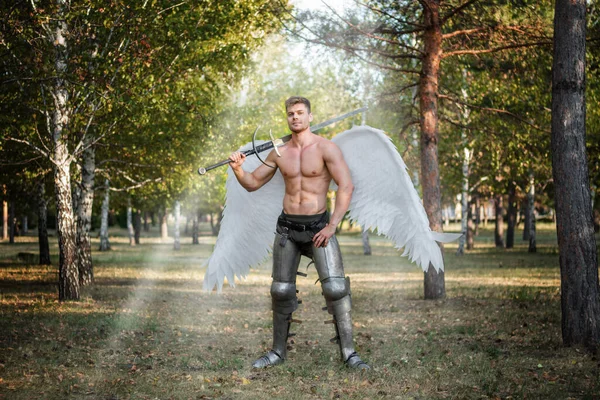 Angel warrior, a bodybuilder in plate armor on his legs with wings behind his back, stands in the beam of light and holds a two-handed sword in his hands.