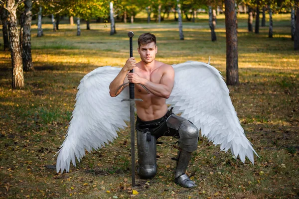 Portrait of a warrior angel with wings behind his back, with a naked torso and in plate armor on his feet, sitting on one knee and holding on to a large sword.