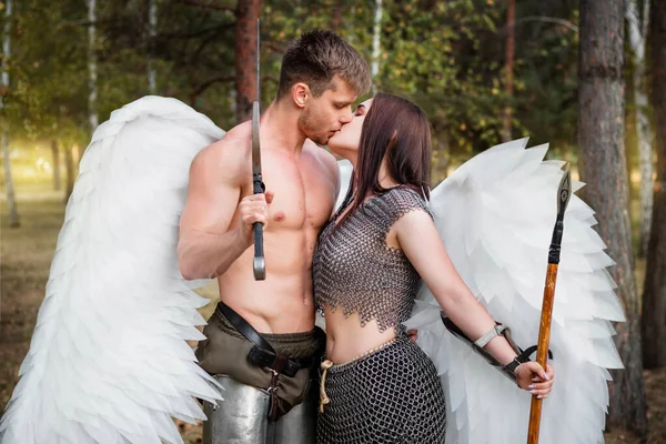 A loving couple of warrior angels kissing. Athletic man and woman in armor with weapons in their hands and white wings behind their backs against the backdrop of the park.