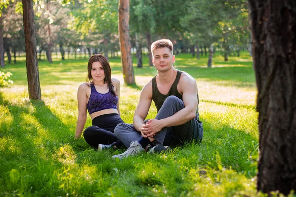 A young, athletic couple, a guy and a girl are sitting nearby in a park on a lawn.