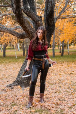 A beautiful girl in a shirt, corset and tight trousers with a saber and a pistol in her hands against the background of an autumn park in the evening. Adventurer woman concept. clipart