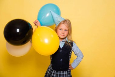 Kafasında şenlik şapkası olan okul üniformalı bir kız elinde sarı bir arka planda izole edilmiş balonlar tutuyor..