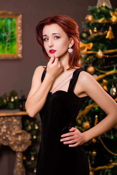 クリスマスツリーを背景にポーズをとった黒のイブニングドレスの魅力的な若い女性の肖像 — ストック写真