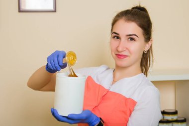 Üniformalı ve mavi lastik eldivenli bir kız saç dökülmesi için elinde sıvı şeker tutuyor. İş yerindeki bir güzellik salonunda şeker tüy dökücü uzmanı..