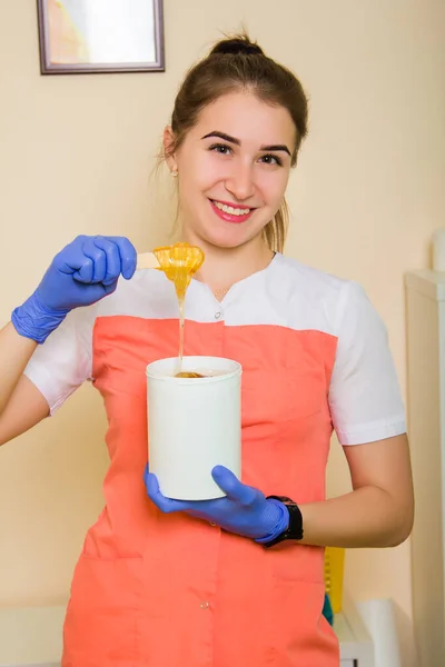 A girl in uniform and blue rubber gloves holds liquid sugar in her hands for hair depilation. Master of sugar depilation in a beauty salon at the workplace.