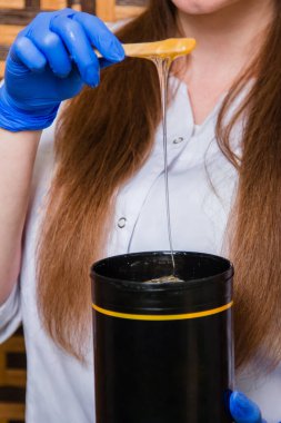 Çubuğun üzerindeki yakın çekim likit şeker tüy dökücü ustasının ellerinde siyah bir kavanoza akar. Beyaz önlüklü ve mavi eldivenli bir kız saç dökülmesi için bir alet tutuyor..