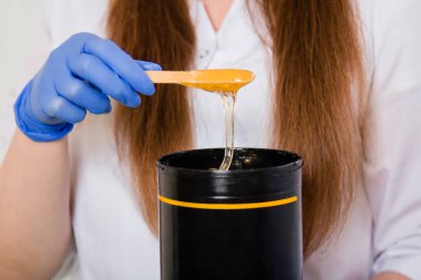 Çubuğun üzerindeki yakın çekim likit şeker tüy dökücü ustasının ellerinde siyah bir kavanoza akar. Beyaz önlüklü ve mavi eldivenli bir kız saç dökülmesi için bir alet tutuyor..