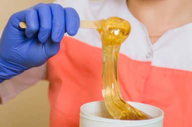 Çubuğun üzerindeki yakın çekim likit şeker tüy dökücü ustasının ellerinde siyah bir kavanoza akar. Üniformalı ve mavi eldivenli bir kadının elinde saç dökülmesi için bir alet var..