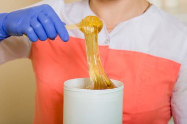 Çubuğun üzerindeki yakın çekim likit şeker tüy dökücü ustasının ellerinde siyah bir kavanoza akar. Üniformalı ve mavi eldivenli bir kadının elinde saç dökülmesi için bir alet var..