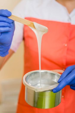 Sıvı sıcak balmumunun yakın plan bir tüy dökücü ustasının ellerinde bir kavanoza akması. Üniformalı ve mavi eldivenli bir kadın saç aldırma macunu tutuyor..