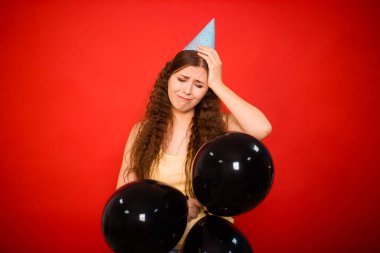 Mutsuz, başarısız doğum günü konsepti. Kafasında şenlik şapkası olan genç bir kadın ellerinde kırmızı arka planda izole edilmiş siyah balonlar tutuyor..