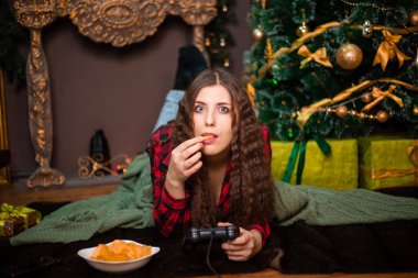 Genç ve duygusal bir kadın, uzaktan kumandayı elinde tutarken video oyunları oynuyor. Noel ağacının yanındaki kürk halıda yatıyor..