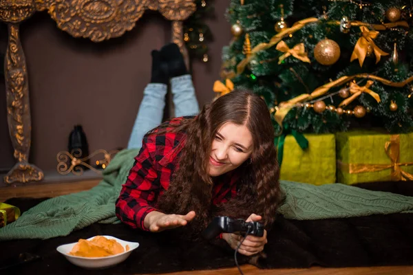 クリスマスツリーの近くの毛皮のカーペットに横たわって 彼女の手のコンソールからリモートコントロールを保持しながら 感情的な女性はビデオゲームをプレイ — ストック写真