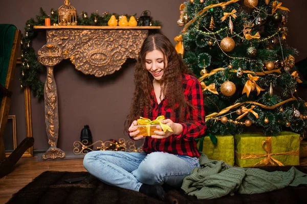 クリスマスツリーを背景に毛布のカーペットに座っている彼女の手に贈り物を持つ魅力的な若い女性 — ストック写真