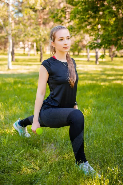 An attractive girl in a sports uniform makes a lunge exercise to stretch the muscles of the legs with dumbbells in her hands. The concept of independent fitness training in nature.