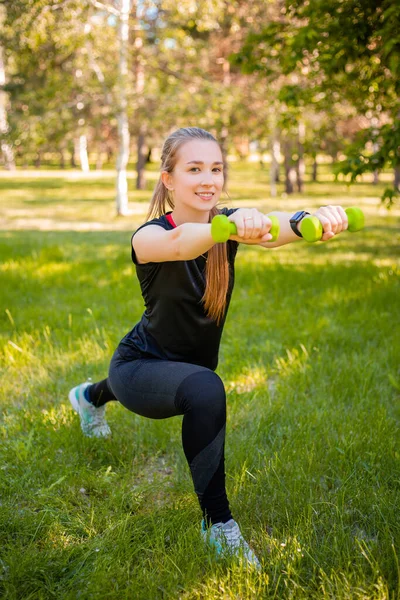 An attractive girl in a sports uniform makes a lunge exercise to stretch the muscles of the legs with dumbbells in her hands. The concept of independent fitness training in nature.