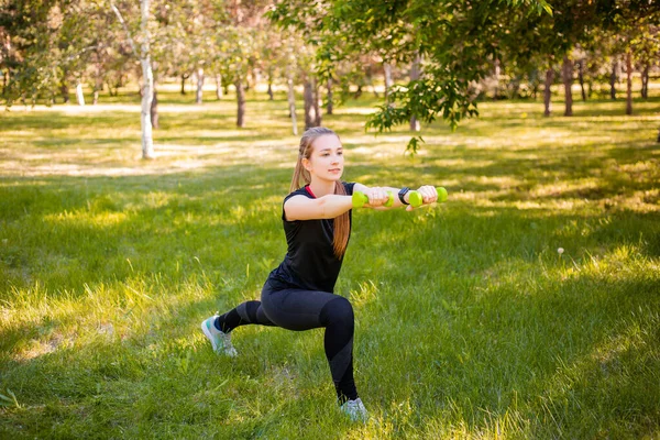 An attractive girl in a sports uniform makes a lunge exercise to stretch the muscles of the legs with dumbbells in her hands. The concept of independent fitness training in nature.