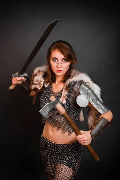 Barbarian woman warrior in chainmail armor with bracers, armor shoulder pad, polar fox fur on her shoulders stands in a fighting stance with an ax and a sword in her hands against a dark background.