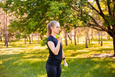 Dambıllarla egzersiz yapan genç bir kadın. Sağlıklı yaşam tarzı kavramı. Parkta spor eğitimi..