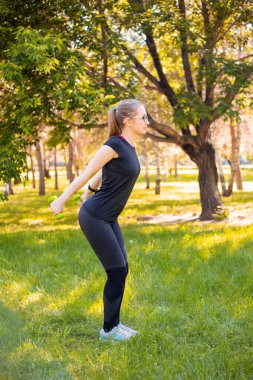 Dambıllarla egzersiz yapan genç bir kadın. Sağlıklı yaşam tarzı kavramı. Parkta spor eğitimi..