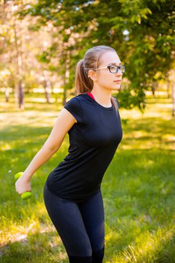 Dambıllarla egzersiz yapan genç bir kadın. Sağlıklı yaşam tarzı kavramı. Parkta spor eğitimi..