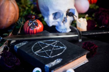 Üzerinde pentagram olan siyah bir kitaba odaklan. Siyah bir masanın üzerinde duran bir kafatası ve balkabağı. Cadılar Bayramı sihirli ritüel kavramı.
