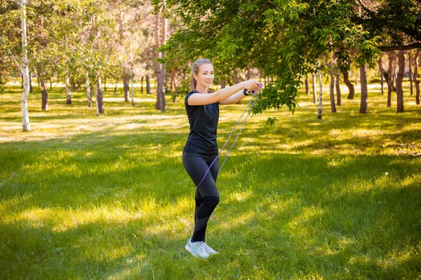 A young woman with a skipping rope in her hands stands on the lawn in a summer park. The concept of a healthy lifestyle and outdoor exercise.