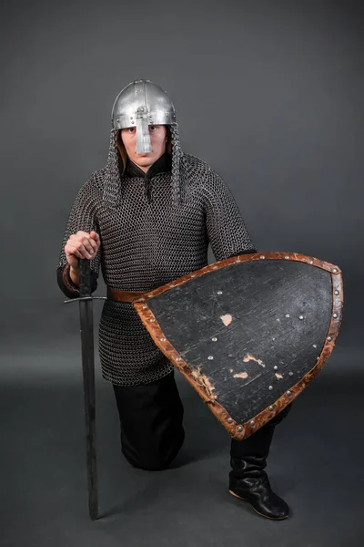 Portrait of a medieval warrior of the late viking era and the beginning of the crusades sitting on his knee. Knight in chain mail and helmet armed with shield and sword isolated on dark background.