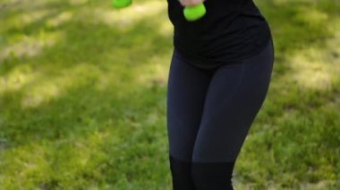 Aşağıdan yukarıya doğru düzgün bir kamera hareketi. İnce, atletik bir kız halterlerle egzersiz yapar. Yaz parkında spor.