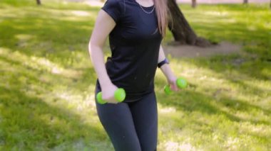 Aşağıdan yukarıya doğru düzgün bir kamera hareketi. İnce, atletik bir kız halterlerle egzersiz yapar. Yaz parkında spor.