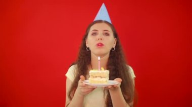 Mumları söndürme ve doğum günü dileği dileme girişimi başarısız oldu. Şenlik şapkalı genç kadın elinde bir dilim pasta ve kırmızı arka planda izole edilmiş bir mum tutuyor..