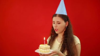 Şenlik şapkalı genç kadın elinde bir dilim pasta ve kırmızı arka planda izole edilmiş bir mum tutan bir tabak tutuyor. Bir mum üflüyorum ve doğum günü dileği tutuyorum..