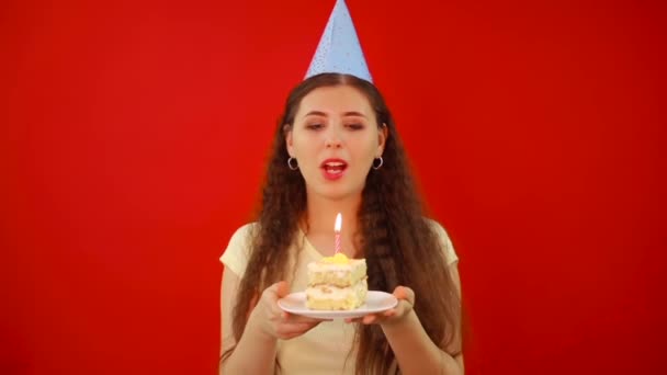 お祝いのキャップの若い女性は ケーキのスライスと赤の背景に隔離された1つのライトアップされたキャンドルを持つプレートを保持します ろうそくを吹いて誕生日の願いを叶えましょう — ストック動画
